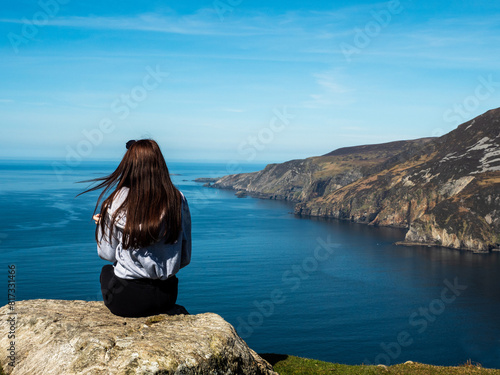 Teenager girl on a trip to Slieve League Cliff, Ireland. Stunning nature scenery with cliff, ocean and clean blue sky. Travel and tourism concept. Model posing in nature scene. Sunny day.