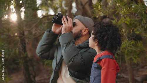 A bearded man with binoculars and a child in a forest engrossed in birdwatching together, surrounded by trees. photo