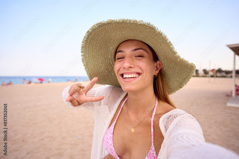 Selfie of generation z Caucasian smiling woman wearing straw hat making hand sign of peace and taking photo standing on beach. Attractive young blonde girl enjoying summer travel posing happy outdoors