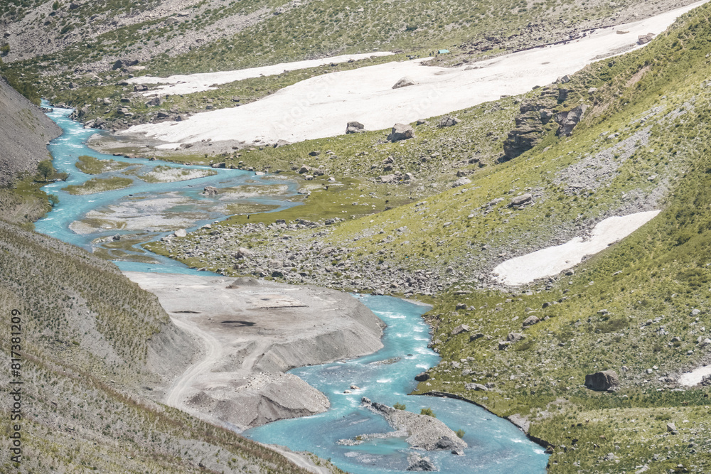 A blue mountain river flows in a mountain valley through the snow past green meadows, a mountain landscape with a river in the mountains of Tajikistan