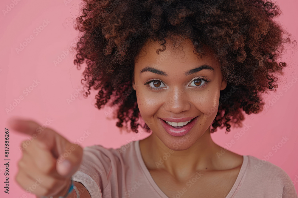 Photo of a happy black woman with afro hair wearing a casual t-shirt pointing his index finger at the camera on a pink background