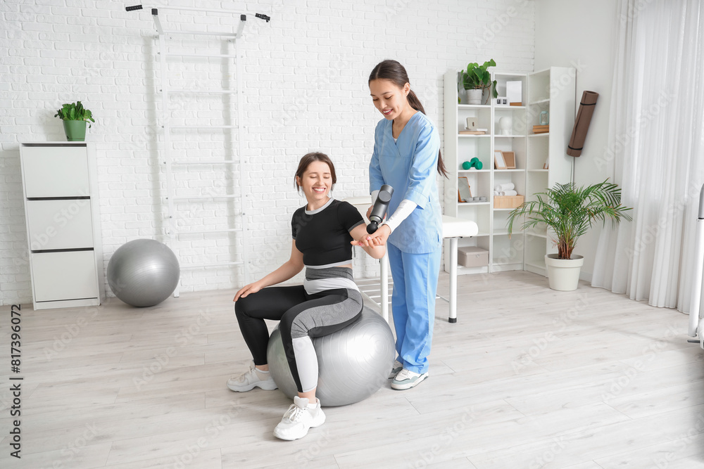 Female Asian physiotherapist massaging young woman's arm with percussive massager on fitball in rehabilitation center Female Asian physiotherapist massaging young woman's arm with percussive massager on fitball in rehabilitation center
