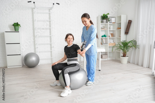 Female Asian physiotherapist massaging young woman's arm with percussive massager on fitball in rehabilitation center