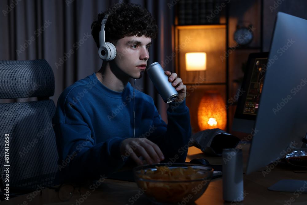 Young man with energy drink and headphones playing video game at wooden desk indoors Young man with energy drink and headphones playing video game at wooden desk indoors