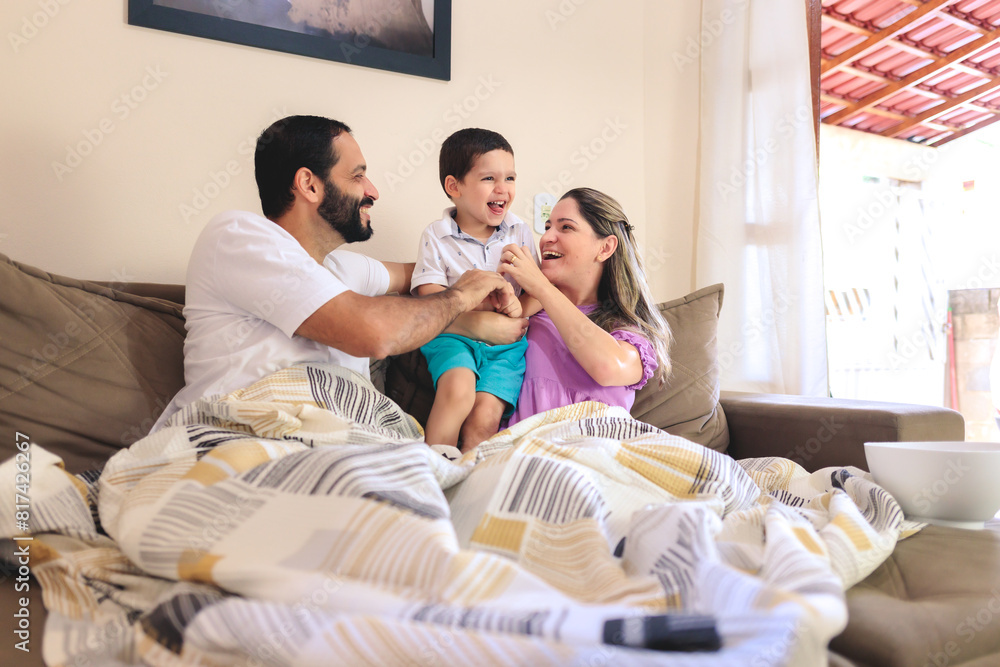 Happy family moment: parents and young son laughing together on the couch, sharing tickles and smiles while watching TV. Perfect for showcasing joyful, authentic family interactions at home. Happy family moment: parents and young son laughing together on the couch, sharing tickles and smiles while watching TV. Perfect for showcasing joyful, authentic family interactions at home.