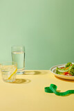 Vertical photo on pastel green background and yellow counter top with blank space in center, surrounded by a measuring tape, a dish of vegetable salad, water and a lemonade. Blank space for design