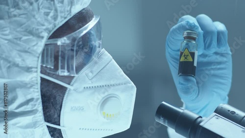 African American laboratory worker in protective overall suit, face mask, safety glasses and mask examining contagious blood sample in test tube with biohazard sticker. Close-up shot photo