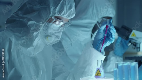Female chemist wearing protective coverall suit, mask, glasses and gloves using pipette and dripping purple reagent into flask with transparent biohazard liquid during experiment in laboratory