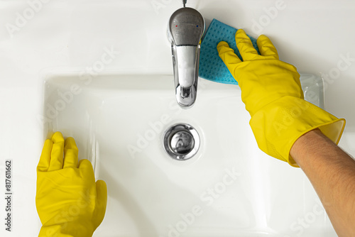 Hygiene concept aerial close up view of female hands wearing yellow protective rubber gloves cleaning a white modern ceramic sink around chrome tap or faucet and fittings using a blue sponge cloth