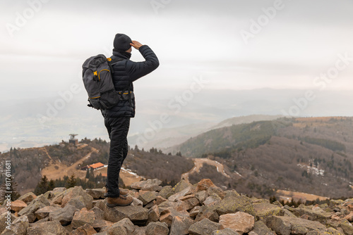 A male hiker stands on top of a rocky outcrop and looks out over the landscape shielding his eyes with his hand; a hike with a large backpack stand on a hill and admires the view on a winter day.