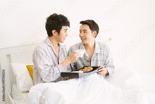 Cute Asian gay couple having breakfast in bed in the bedroom. Happy gay couple wearing pajamas having breakfast in morning in bedroom, Looking into each others eyes and Smiling