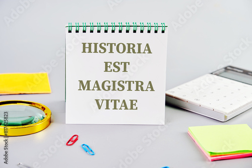 Historia est vitae magistra (History is the tutor of life) Latin phrase written on a notepad on a gray background next to office supplies photo