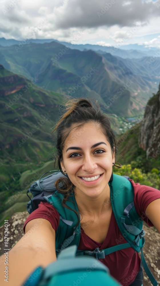 A woman is smiling and taking a selfie on a mountain. She is wearing a green backpack and a red shirt