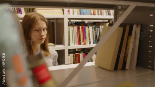 Young female student taking book from shelf and reading it in college library