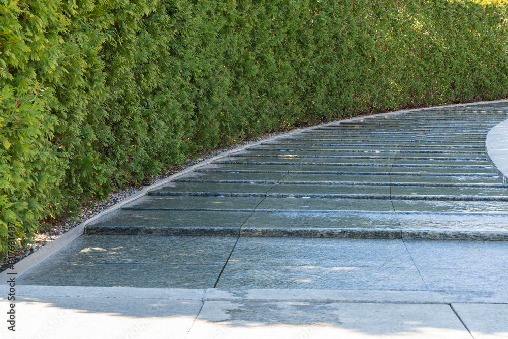 Obraz premium Artificial swift river with granite banks runs around evergreen hedge with rest area. Public landscape 'Galitsky park' for relaxation and walking in sunny spring 2024