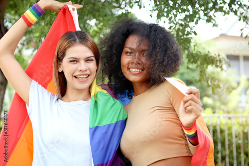 Happy smiling homosexual lesbian couple holding rainbow flag. Beautiful woman and her African curly hair girlfriend celebrate pride month together, romantic LGBT lover and gay pride movement concept