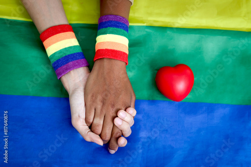 Hands of homosexual lesbian couple with rainbow wristband holding hand each other on rainbow flag with red heart, Diverse lover hands, celebration of LGBT gay pride month.