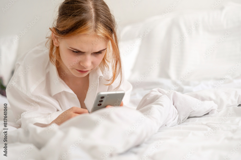 Serious bored young girl with gloomy face expression looking at smartphone screen, teenager using mobile phone laying in bed