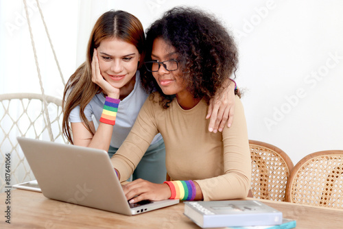 Happy homosexual lesbian couple with rainbow flag wristband using laptop computer together. Beautiful woman and African girlfriend search for pride celebration gifts online. Romantic LGBT lover family
