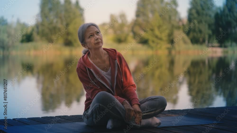 Calm senior woman sits on fitness mat on wooden pier, preparing for yoga Calm senior woman sits on fitness mat on wooden pier, preparing for yoga
