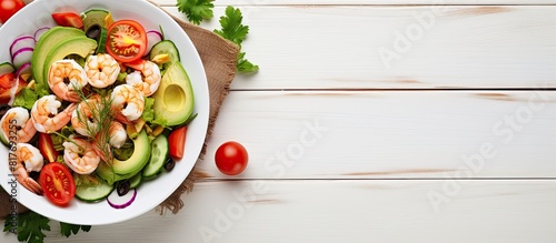 Diet concept featuring an avocado salad topped with shrimp and vegetables The white wooden background provides copy space image