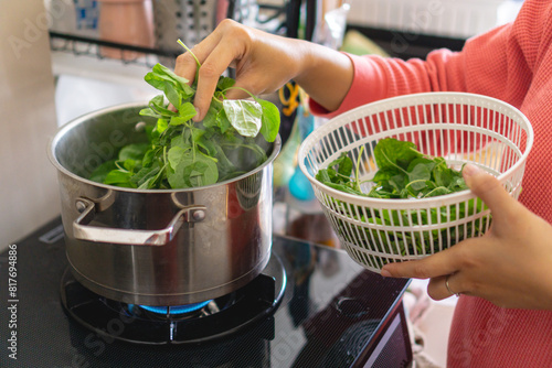 A woman is blanching or cooking spinach in a pot on a stove. She is holding a basket of spinach in her hand, home cooking concept