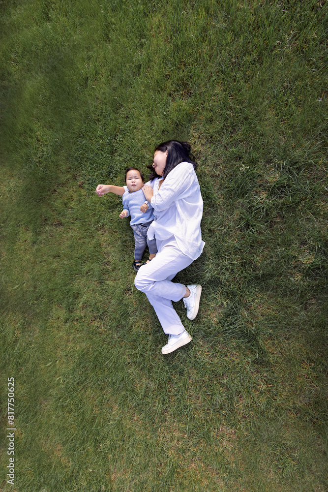 Asian smiling mom and sweet daughter are lying on a green lawn. A charming Korean woman and her cute kid are having fun on the grass.