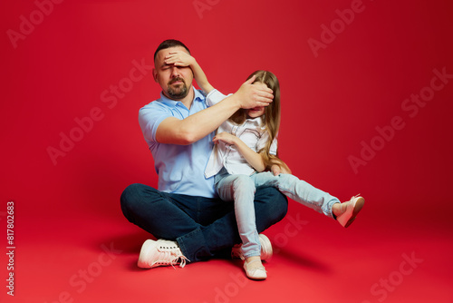 Man sits on floor with young girl, his daughter and they covered eyes to each other against vibrant red studio background. Concept of Father's day. Children's day, Family day, parenthood. Ad