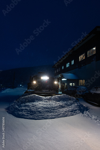 A snowcat, waiting for a group of skiers and snowboarders, clears the snow near the entrance to the hotel early in the morning during a snowfall and illuminates everything with headlights