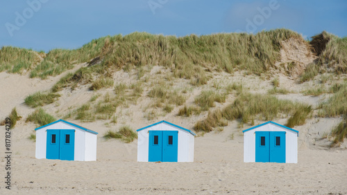 Three colorful beach huts stand in a row on the sandy shores of Texel, Netherlands, under a clear blue sky.