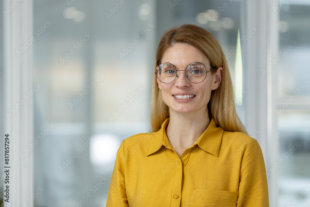 Confident and happy female professional in a yellow shirt and glasses smiling in a modern office environment. Confident and happy female professional in a yellow shirt and glasses smiling in a modern office environment.