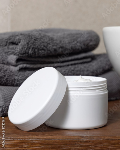 White cosmetic jar near grey folded towels and basin on wooden countertop in bath, mockup