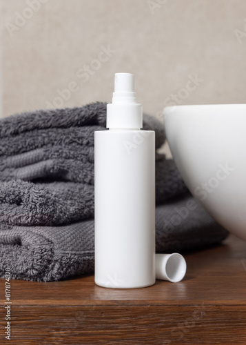 White cosmetic bottle near grey folded towels and basin on wooden countertop in bath, mockup