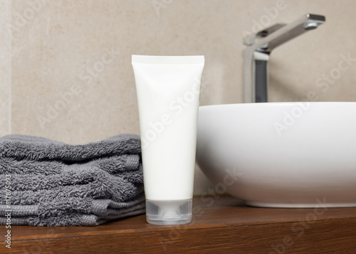 White cosmetic tube near grey folded towels and basin on wooden countertop in bath, mockup