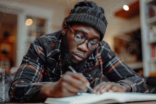A young man with glasses and a beanie, intensely focused on writing in a notebook. The cozy indoor setting creates a warm and studious atmosphere.