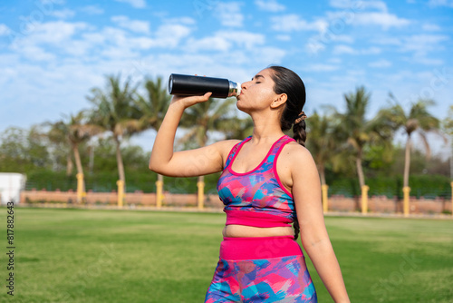 Fit Young indian woman drinking water after outdoor workout or training. young female athlete relax, resting after exercise in park or garden. break, dehydration concept. .