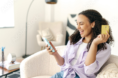 Cheerful latino or brazilian woman, holding banking card and mobile phone to buy order prepay goods online at web store via special app while sitting at home in living room on sofa, smiling, rejoicing