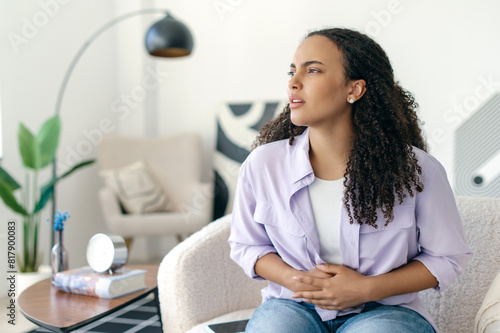 Unhappy sick young curly haired brazilian or hispanic woman in casual clothes, sitting on couch in a living room at home, touching belly, suffering from period cramps, stomachache, feels pain
