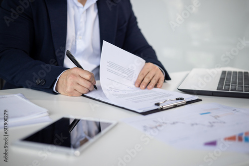 Asian mature man in a business suit signing important documents at an office desk with a laptop, tablet, and papers.