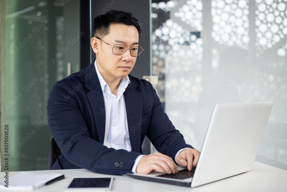 Asian mature man in a business suit working on a laptop computer in a modern office setting, focusing on his task. Asian mature man in a business suit working on a laptop computer in a modern office setting, focusing on his task.