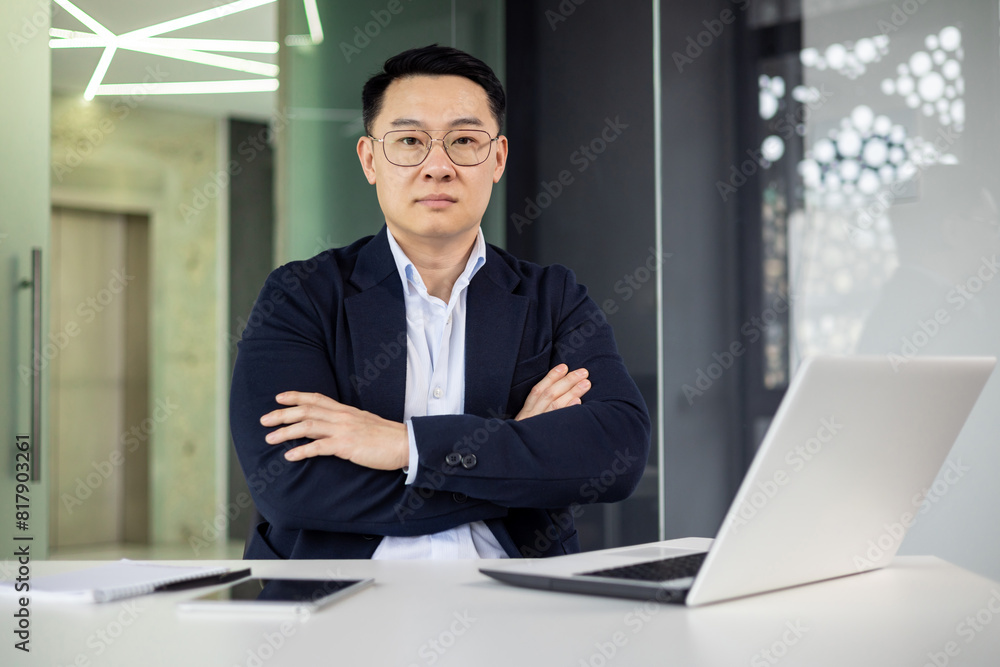 Confident asian mature man in formal attire with arms crossed, sitting at a desk in modern office with laptop and notepad. Confident asian mature man in formal attire with arms crossed, sitting at a desk in modern office with laptop and notepad.