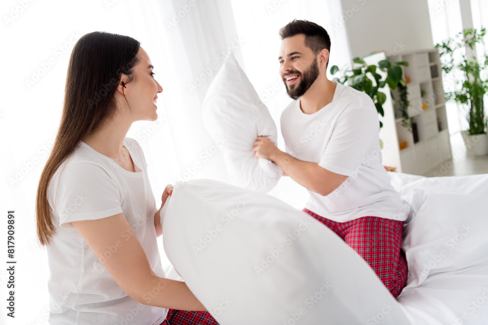 Fototapeta premium Photo of childish fooling couple wear white t-shirts pillow fighting indoors apartment bedroom
