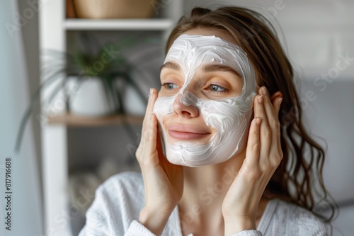 A woman enjoying a skincare routine is shown in a home setting with a creamy facial mask applied to her face while wearing a white robe.