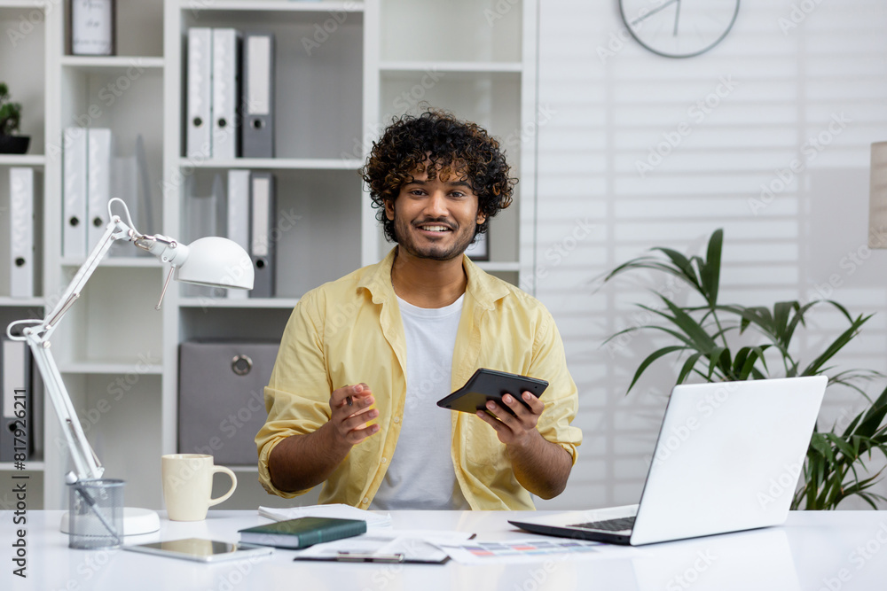 Young man in yellow shirt working at desk with laptop, calculator, and paperwork in modern office environment with plants and shelves.