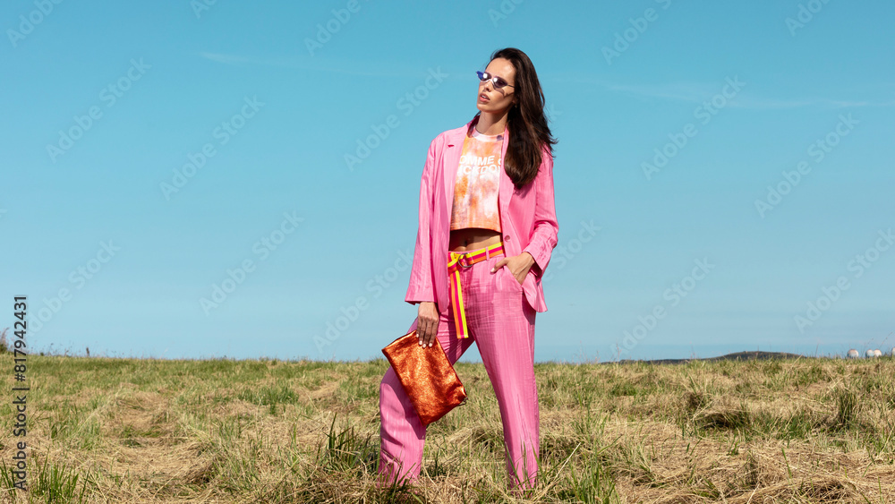 model fashion trend  women. Model with pink jacket and pants suit and orange handbag with sunglasses in the sun with light blue sky background, spring summer fashion editorial