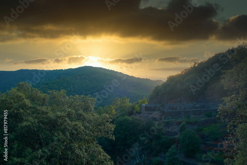 A breathtaking view of a sunset over a verdant landscape featuring rolling hills and terraced greenery. The golden sunlight breaks through the clouds, casting a warm glow over the lush vegetation. The