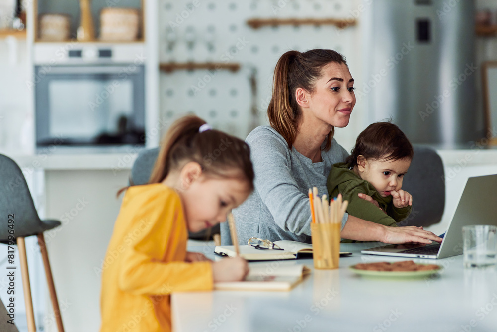 An overworked mother working on her laptop while taking care of her children An overworked mother working on her laptop while taking care of her children