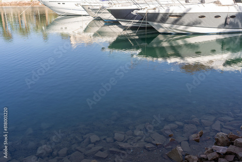 Clear water reflects luxury yachts docked in a serene marina, with rocks visible at the bottom. A peaceful scene showcasing marine leisure.