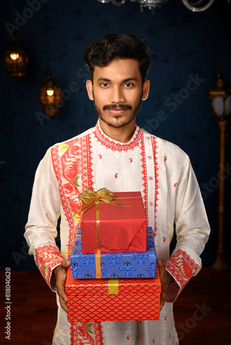 Portrait of a young and handsome man, clad in a traditional kurta pyjama, holding gift boxes in hands under studio lighting. Indian fashion, events and lifestyle. photo