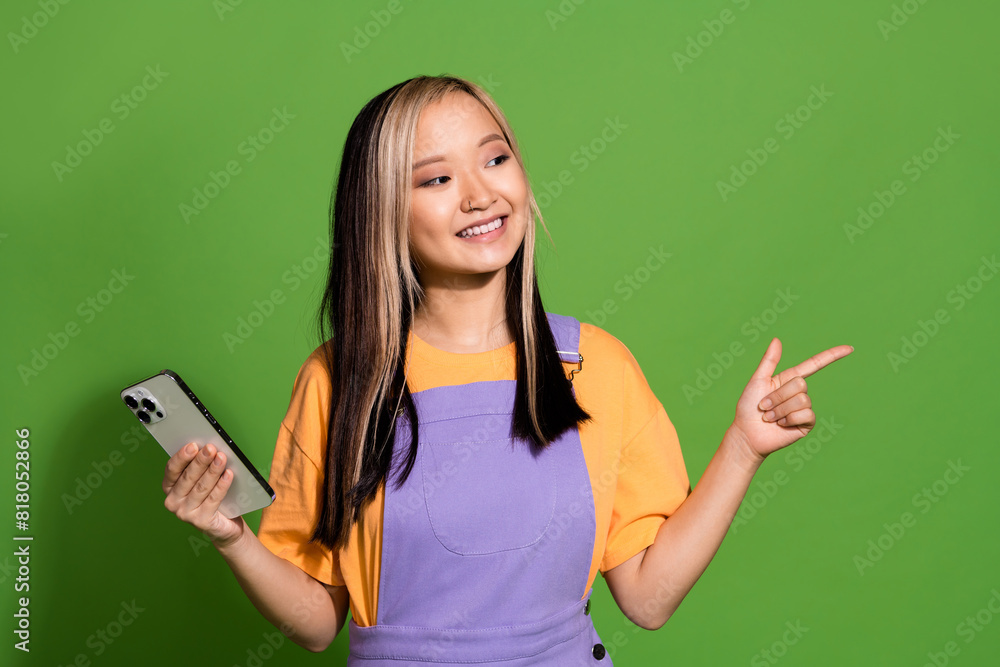 Photo of lovely young lady hold gadget look point empty space dressed stylish violet yellow garment isolated on green color background Photo of lovely young lady hold gadget look point empty space dressed stylish violet yellow garment isolated on green color background